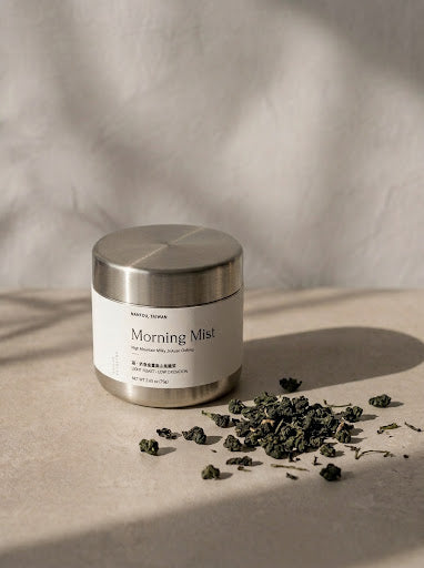 A jar of MISTEL CLEAN Morning Mist skincare product with a brushed silver lid and white label, styled on a neutral beige surface with scattered dried green tea leaves under soft, dappled shadows.
