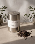A stainless steel tea tin labeled 'Golden Hour' sits on a light surface with loose dried tea leaves beside it, styled with soft shadows and small plants in the background.