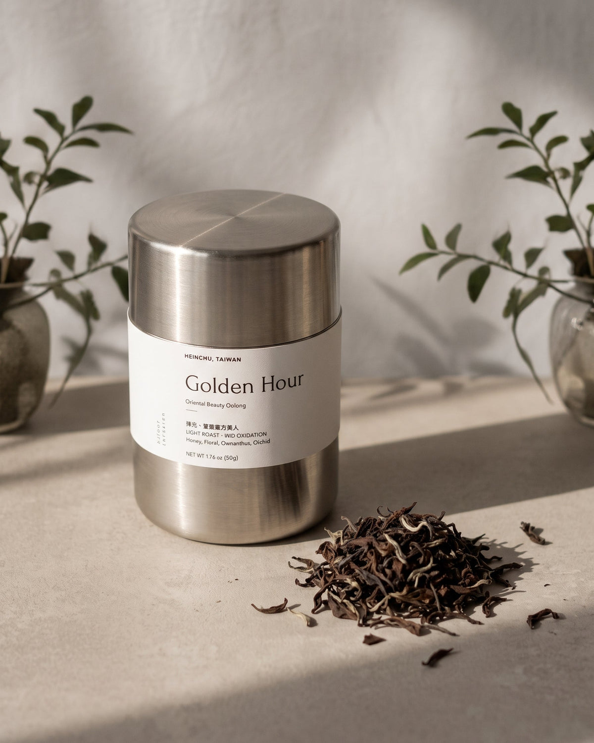 A stainless steel tea tin labeled 'Golden Hour' sits on a light surface with loose dried tea leaves beside it, styled with soft shadows and small plants in the background.