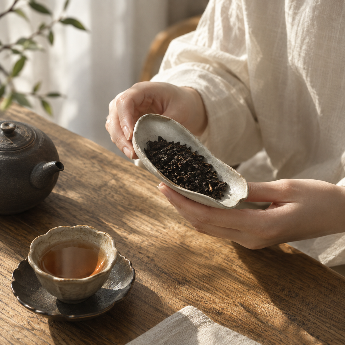 A person in a white linen shirt holds a small speckled ceramic scoop filled with loose-leaf tea over a wooden table. A teapot and a cup of tea sit nearby in a warm, softly lit setting.