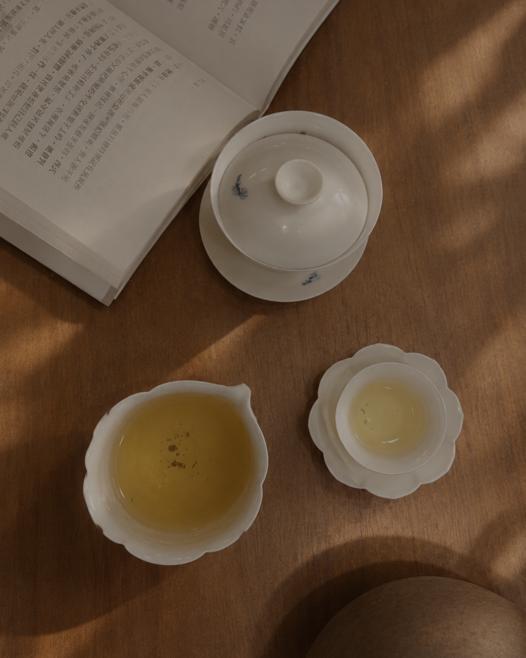 White ceramic tea set with a gaiwan, teapot, and small cups arranged on a wooden table next to an open book, with tea inside the cups.